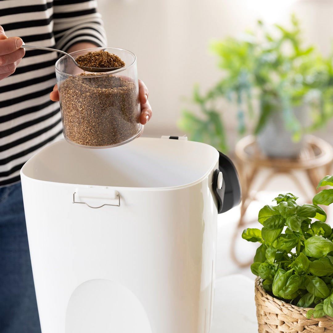 Person adding granulate to a white Bokashi bin with plants in the background